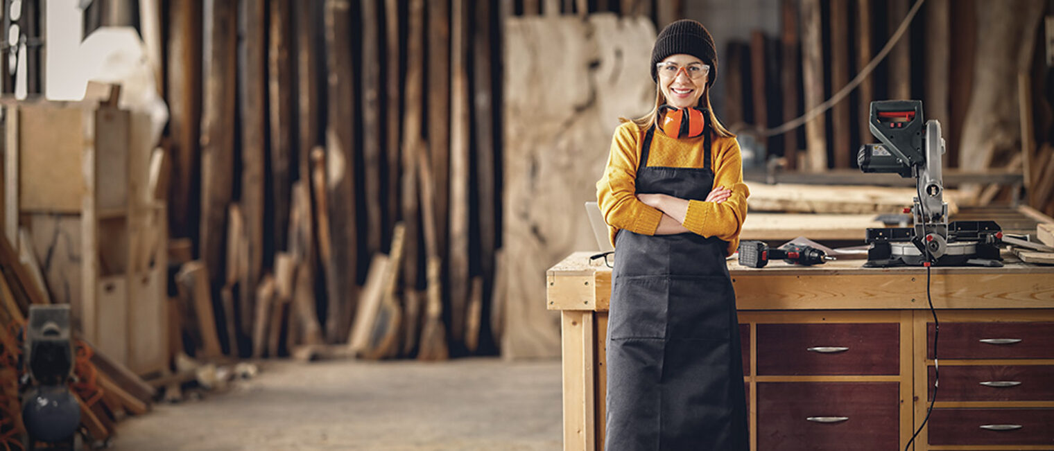 Full body of smiling confident young female joiner in apron standing near workbench and looking at camera friendly while working in craft workshop Schlagwort(e): carpenter, woman, joiner, workshop, artisan, craft, work, woodworker, cheerful, small business, craftswoman, carpentry, professional, happy, smile, female, young, confident, occupation, job, skill, craftsmanship, positive, owner, lifestyle, joinery, manufacture, handicraft, production, handmade, master, maker, labor, worker, indoors, service, arms crossed, full length, full body, looking at camera, blurred background, yellow sweater, girl, young, workshop