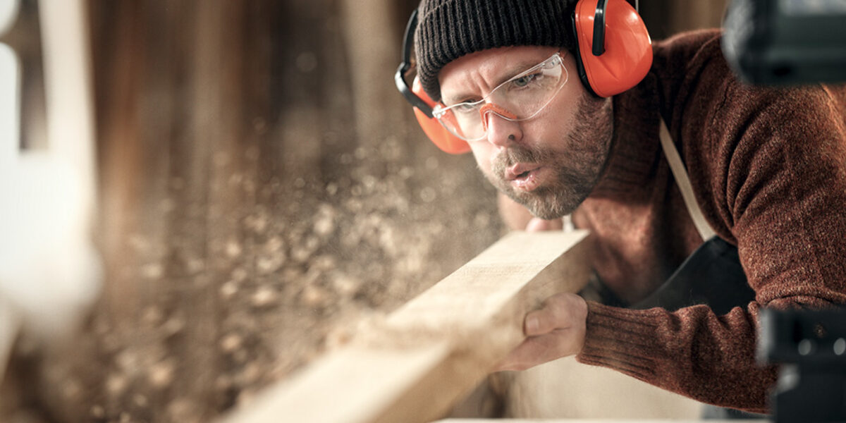 Adult male woodworker in protective goggles and headphones blowing sawdust from wooden detail while working in carpentry workshop Schlagwort(e): carpenter, woodwork, joiner, blow, sawdust, wood, protect, goggles, headphones, work, workshop, man, craftsman, artisan, male, adult, woodworker, carpentry, skill, small business, craft, safety, professional, occupation, job, craftsmanship, joinery, manual, handmade, handicraft, process, handwork, busy, maker, labor, plank, dust, wooden, create, worker, timber, lumber, indoors, blur, selective focus, crop