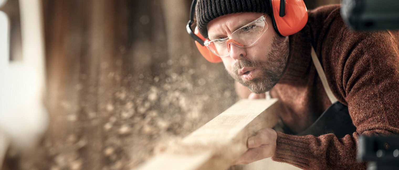 Adult male woodworker in protective goggles and headphones blowing sawdust from wooden detail while working in carpentry workshop Schlagwort(e): carpenter, woodwork, joiner, blow, sawdust, wood, protect, goggles, headphones, work, workshop, man, craftsman, artisan, male, adult, woodworker, carpentry, skill, small business, craft, safety, professional, occupation, job, craftsmanship, joinery, manual, handmade, handicraft, process, handwork, busy, maker, labor, plank, dust, wooden, create, worker, timber, lumber, indoors, blur, selective focus, crop