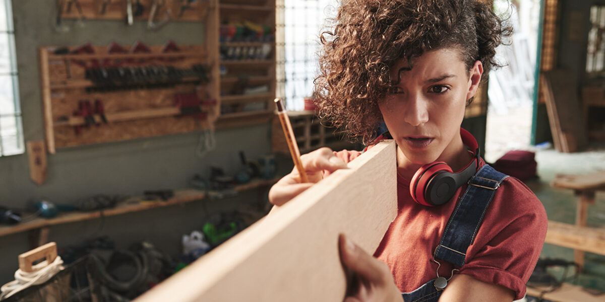 Young concentrated female carpenter with curly hair holding wooden plank and estimating its length before sawing Schlagwort(e): woman, female, young, pretty, curly, denim, overall, concentrated, focused, carpenter, carpentry, joinery, timber, wood, wooden, plank, measuring, measurement, length, eye, sight, tool, skill, manual, equipment, professional, craft, craftsman, handyman, occupation, working, woodworking, workshop, lumber