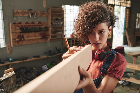 Young concentrated female carpenter with curly hair holding wooden plank and estimating its length before sawing Schlagwort(e): woman, female, young, pretty, curly, denim, overall, concentrated, focused, carpenter, carpentry, joinery, timber, wood, wooden, plank, measuring, measurement, length, eye, sight, tool, skill, manual, equipment, professional, craft, craftsman, handyman, occupation, working, woodworking, workshop, lumber